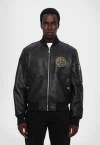 Giubbotto bomber in pelle nera con polsini e orlo a coste, caratterizzato da una patch con logo dorato sul petto e una tasca con zip sulla manica.