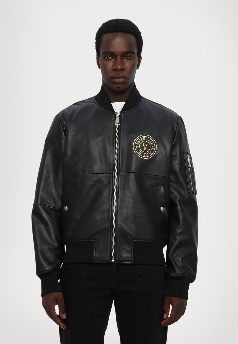 Giubbotto bomber in pelle nera con polsini e orlo a coste, caratterizzato da una patch con logo dorato sul petto e una tasca con zip sulla manica.