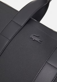 Gros plan sur un sac en cuir noir texturé et lisse avec des poignées cousues et un petit logo crocodile embossé.