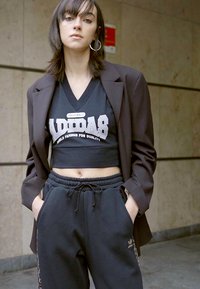 Fekete, rövid Adidas top fehér logóval, amely fekete melegítőnadrággal van párosítva, logó részletekkel, és egy oversized barna blézert visel.