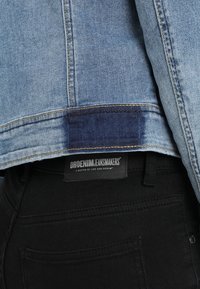 Světle modrá denimová bunda s tmavě modrým textilním záplatou na lemu, spárovaná s černými džíny. Viditelný štítek s nápisem "DRDENIM JEANMAKERS".