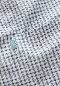 Biely textil s ružovo-zeleným kockovaným vzorom, s malou zelenou značkou s bielym logom hlavy jeleňa.