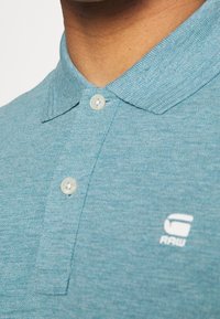 Polo bleu sarcelle avec une finition texturée, doté d'une patte à deux boutons et d'un logo blanc sur le côté gauche de la poitrine.
