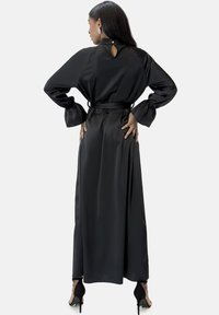 Zwarte satijnen lange jurk met een keyhole-rug, taille met ceintuur en brede mouwen. Glanzende textuur en gladde afwerking, enkelhoge lengte.