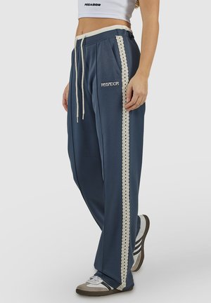 Woman wearing navy blue sweatpants with cream lace stripe, drawstring waist, and white crop top, standing with one foot forward.