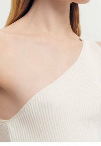 Femme portant un haut blanc à côtes, à une épaule, laissant apparaître la clavicule et le cou, avec des cheveux longs, lisses et châtain clair.