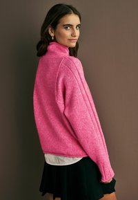 Maglione oversize rosa brillante con colletto alto, lavorazione a maglia texturizzata e polsini a coste, abbinato a una camicia bianca e una gonna nera.