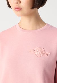 Vrouw die een lichtroze sweatshirt draagt met een verhoogd geborduurd GANT-logo op de borst, geknipt op de hals en schouders.