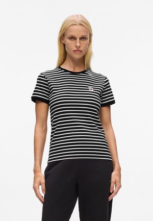 Blonde woman wearing a black and white striped short-sleeve shirt with a small embroidered logo on the chest and black pants.
