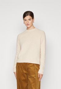 Beiger Strickpullover mit Rundhalsausschnitt und gerippten Bündchen, kombiniert mit braunen Cordhosen. Weicher Stoff, lockere Passform und kurze Länge.