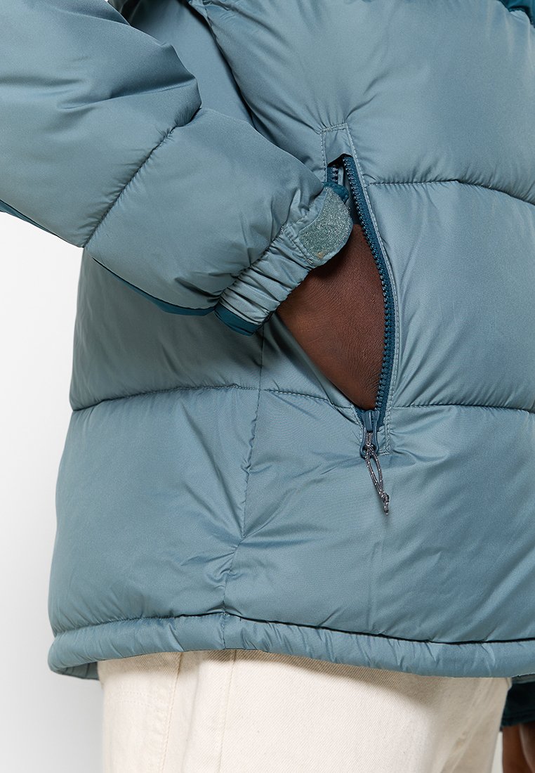 Giacca puffer azzurro chiaro con tasca con zip, polsini elastici e texture trapuntata. Il tessuto appare liscio e leggermente lucido.