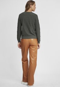 Donkergroene trui met een getextureerd bobbelpatroon, gecombineerd met hoog-getailleerde, glanzende caramelkleurige leren broek. Neutrale sneakers maken de look compleet.