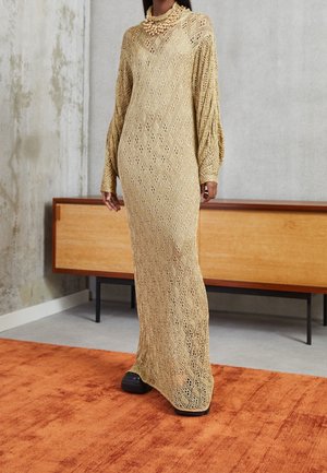 Kobieta w beżowej, długiej, dzianinowej sukience z wzorem w romby, czarnych butach i masywnym naszyjniku z pereł, stojąca w pomieszczeniu.