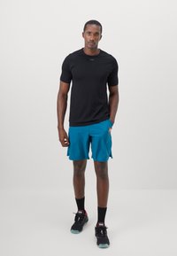 Black athletic t-shirt, teal shorts with side slits, black sneakers with red accents, and black socks, standing on a plain backdrop.