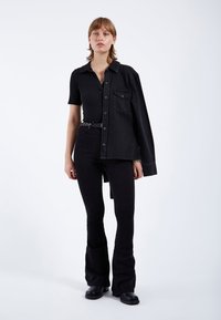 Camisa de mezclilla negra con mangas cortas, abotonada y un bolsillo frontal, combinada con jeans negros acampanados y botas negras gruesas.