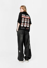 Camiseta oversized negra con un gráfico en rojo y blanco en la parte posterior. Acompañada de unos pantalones holgados de color gris oscuro y zapatos de tono claro.