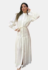 Abito maxi bianco con bottoni, maniche a sbuffo lunghe, vita con laccetto e gonna a strati. Realizzato in tessuto leggero, presenta una chiusura frontale completa con bottoni.
