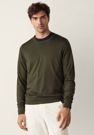 Mann mit lockigem dunklem Haar, der einen olivgrünen Pullover über einem schwarzen Unterhemd und weiße Hosen vor einem schlichten hellen Hintergrund trägt.