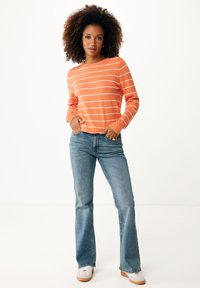 Pomarańczowo-biały sweter w paski z długimi rękawami, zestawiony z jasnoniebieskimi dzwonami i białymi tenisówkami. Model stoi z rękami w kieszeniach.