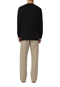 Persona che indossa una camicia nera a maniche lunghe e pantaloni beige, in piedi con la schiena rivolta verso uno sfondo bianco.