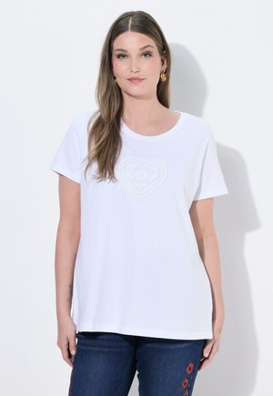 Kvinde med langt brunt hår har en hvid T-shirt med et diskret hjerte-design og mørkeblå jeans med orange blomsterbroderi på.
