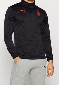 Maglia atletica nera a maniche lunghe con collo a zip, caratterizzata da un logo Puma arancione e un emblema dell'AC Milan arancione, realizzata in materiale liscio e elastico.