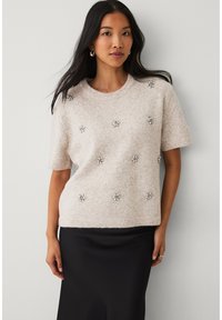 Hellbeiger Strickpullover mit kurzen Ärmeln, verziert mit dekorativen silbernen Blumenapplikationen, die gleichmäßig über die Vorderseite verteilt sind.