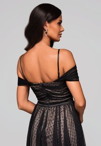 Femme aux longs cheveux noirs portant une robe noire à pois décole épaules et de grandes boucles d'oreilles, faisant face de côté avec son dos visible.