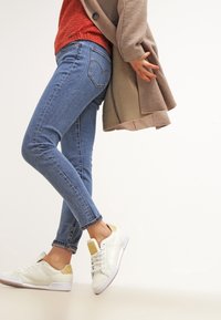 Weiße Sneakers mit gelben Akzenten, kombiniert mit blauen Slim-Fit-Jeans und einer beigen Strickjacke über einem roten Pullover. Fokus auf die Details des lässigen Outfits.