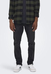 Jeans en denim noir associés à une chemise en flanelle à carreaux verts et noirs. Les chaussures sont blanches avec des accents verts. Design simple et fonctionnel.