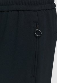 Un pantalon de sport noir présente un tissu texturé, une taille élastique et une poche zippée avec un détail de tirette circulaire.