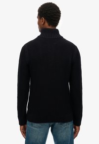 Marineblauer Strickpullover mit hohem Kragen, gerippten Bündchen und strukturiertem Stoff. Kombiniert mit hellblauen Jeans, die ein lässiges Design zeigen.