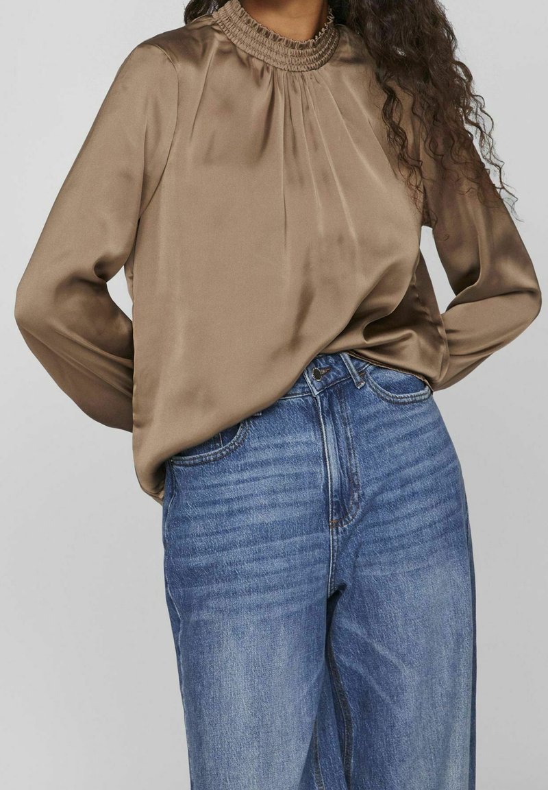 Femme portant un chemisier en satin marron à col montant avec des manches longues rentrée dans un jean bleu taille haute, mains dans le dos.