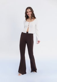 Fehér bordázott crop top gombos részletekkel, barnás, testhezálló, harangszárú nadrággal párosítva. Sima textúra és testhezálló kialakítás, nyitott hasrésszel.