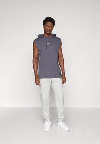 Hõlmikuta tumeda halli kapuutsiga pusa paelade ja logoga, kombineeritud heleda halli dressipükstega. Mudel kannab valgeid tossusid.