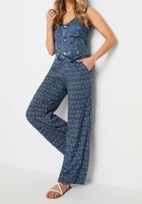 Džínová vesta na knoflíky s dvěma předními kapsami, spojená s širokými modrými kalhotami s květinovým vzorem a bílými sandály.