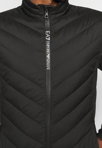 Veste matelassée noire avec col montant et fermeture éclair verticale avec marquage blanc "EA7 Emporio Armani" au centre devant.
