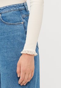 Haut à manches longues en crème, à rayures, avec des poignets en dentelle froncés, associé à un jean en denim bleu avec une poche avant et une coupe classique.