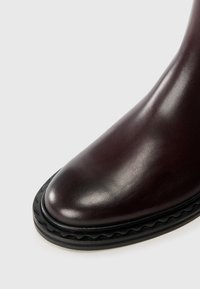 Bottine en cuir bordeaux avec un bout rond, semelle en caoutchouc noir texturé et couture décorative ondulée autour du bord de la semelle.