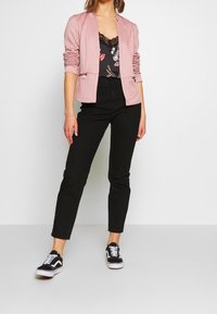 Ljusrosa blazer med skräddarsydd passform, kombinerad med en svart blommig spets topp, svarta slim-fit jeans och svarta sneakers med vita detaljer.