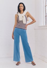 Top senza maniche a righe color melograno e bianco con un maglione crema drappeggiato intorno al collo; abbinato a jeans a gamba larga color azzurro chiaro e sandali neri.