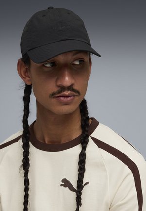 Uomo con capelli lunghi intrecciati che indossa un berretto nero Puma e una maglietta crema Puma con rifiniture marroni, guarda di lato su uno sfondo grigio.