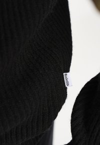 Sort striktrøje med ribbet struktur. Har et lille hvidt mærke med "Re:Designed." Nærbillede af stofdetaljen.