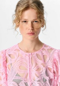 Jeune femme aux cheveux blonds bouclés portant un haut en dentelle rose avec des motifs floraux sur un vêtement gris, faisant face à un fond blanc.