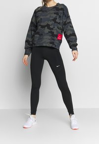 Sweatshirt surdimensionné en motif camouflage dans des verts foncés et noirs, avec un patch logo rouge ; associé à des leggings noirs et des chaussures de sport blanches.