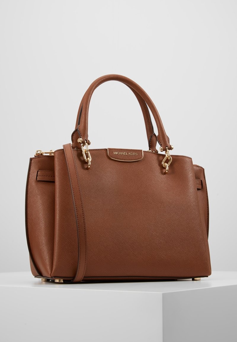 Sac à main en cuir marron Michael Kors avec double poignée, une bandoulière amovible et des ferrures dorées, posé sur une surface blanche.