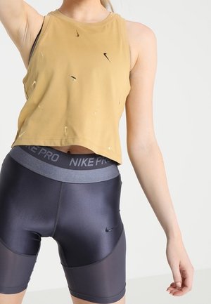 Kobieta w podartym musztardowym topie bez rękawów i czarnych szortach rowerowych Nike Pro, pozująca z uniesioną ręką na jednolitym białym tle.