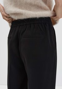 Pantalon noir avec une taille élastique, comprenant deux poches arrière. Le matériau semble lisse avec un léger éclat. Pull partiellement visible au-dessus.