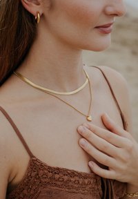 Colier aur stratificat cu un pandantiv în formă de inimă, purtat împreună cu un choker subțire aurit. Modelul poartă un top maro din dantelă, evidențiind tonul natural al pielii.