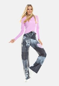 Jauna sieviete smaida, pozējot ar vienu kāju paceltu, valk ājot rozā garām piedurknēm džemperi, plankumainas džinsa bikses un baltus sporta apavus uz balta fona.